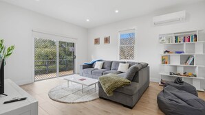 A modern living room with a grey sofa, a coffee table, a bookshelf, and a large window. - Apple Court Retreat – 3 Bedroom – Access to Huon River (Huonville)