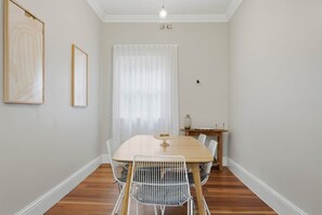 A dining area with a wooden table and white chairs, a window with sheer curtains, and a light fixture above. - Cottage on Quayle - 3 Bedroom in Sandy Bay (Sandy Bay)