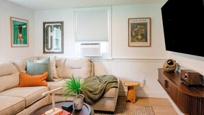 A living room with a sofa, a coffee table, a small stool, and a television. There are framed pictures on the walls and a window with blinds. - Retro Luxe Suite, Downtown Manchester near Hartford Sleeps 4! New! (Manchester)