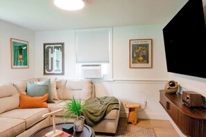 Ein Wohnzimmer mit einer Couch, einem Couchtisch, einem kleinen Hocker und einem Fernseher. An den Wänden hängen Bilder in Rahmen, und ein Fenster mit Jalousien ist zu sehen.