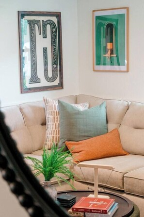 Un salon avec un canapé beige, des coussins à motifs, une petite plante et des œuvres d’art encadrées accrochées au mur.