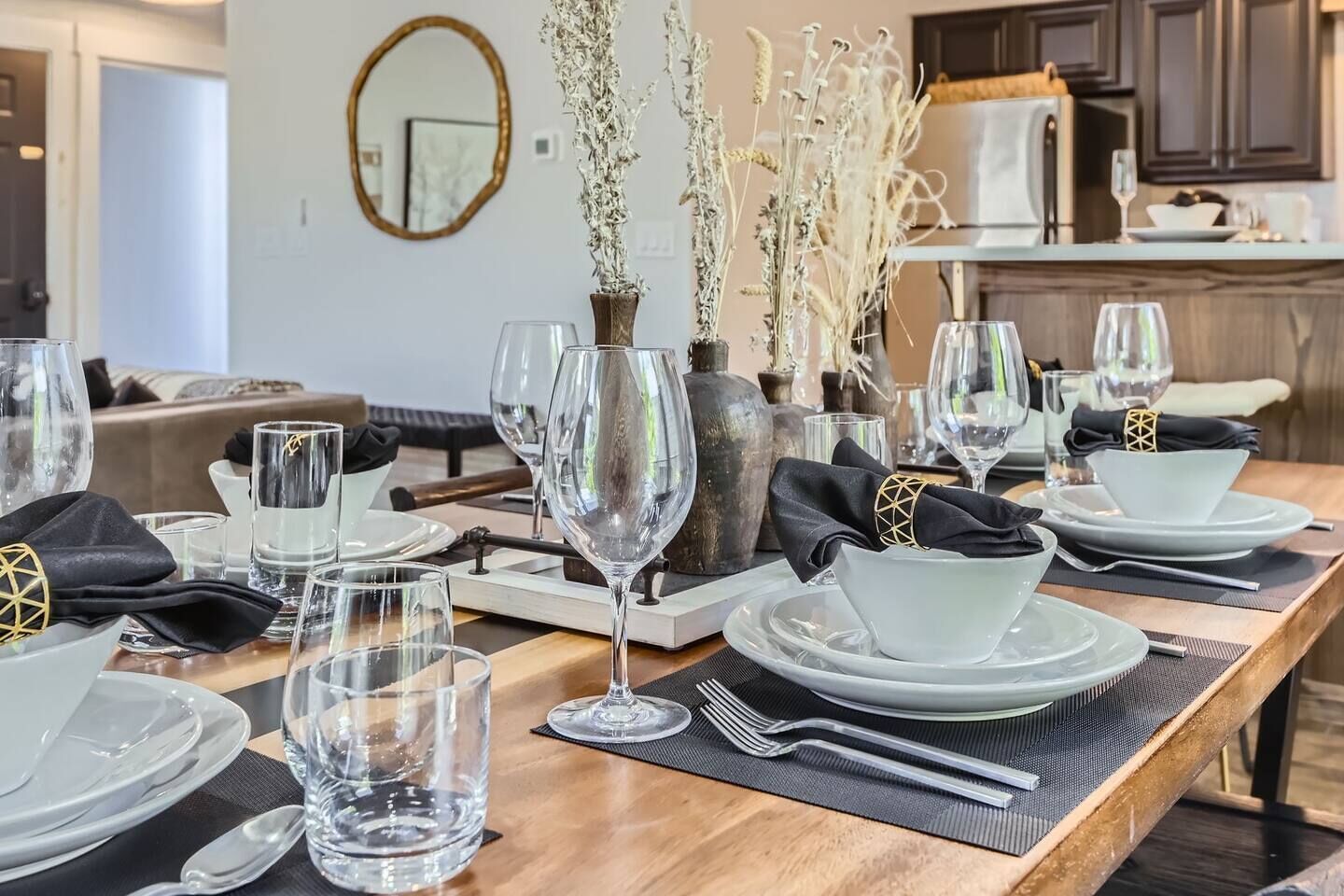 A dining table set with plates, glasses, and cutlery, with a vase of dried flowers in the center.