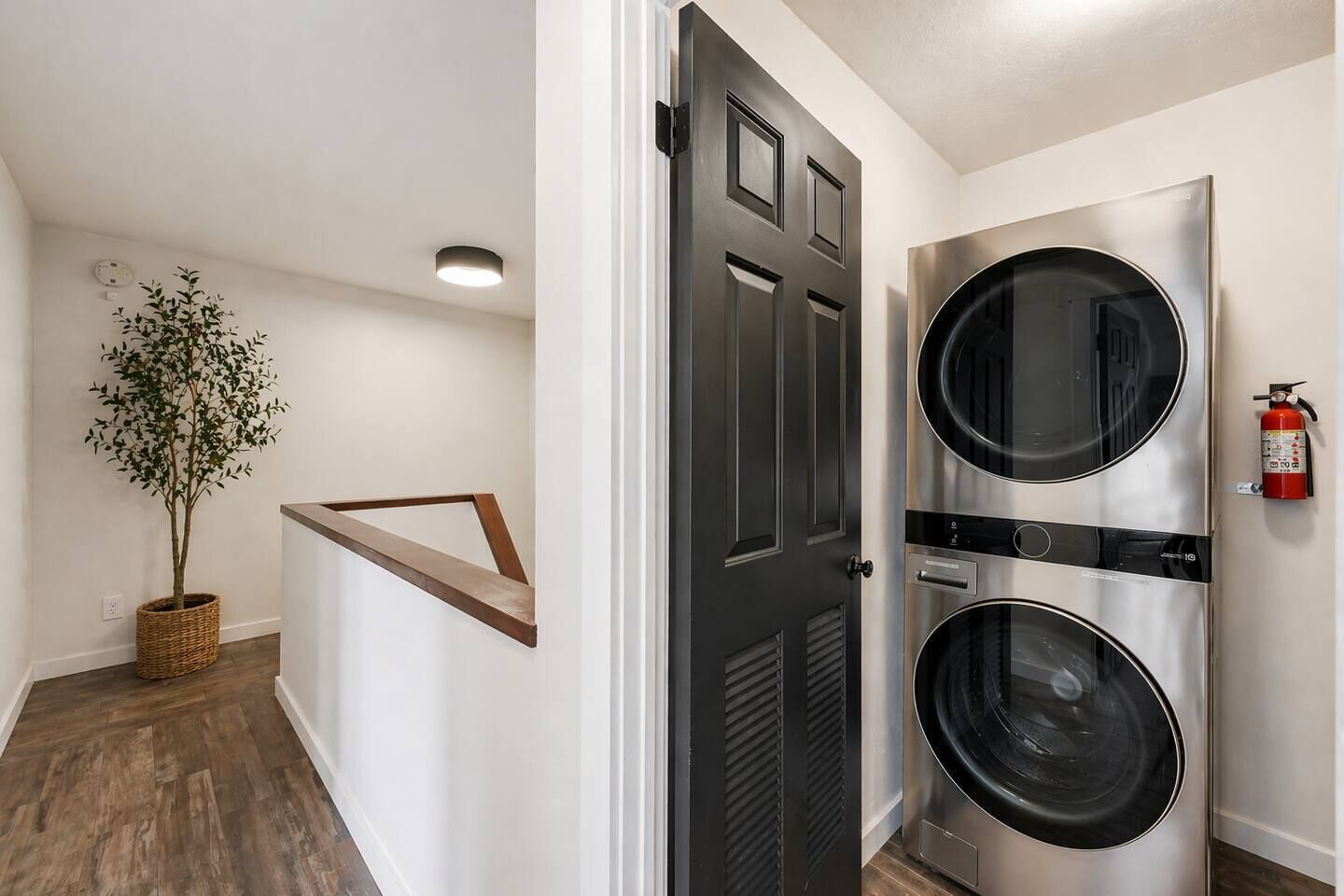 A laundry room with two stacked front-loading washers and dryers, a fire extinguisher on the wall, and a potted plant in the corner.