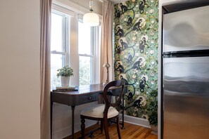 A kitchen with a modern stainless steel refrigerator, a wooden chair, a desk with a plant, and a window with curtains.