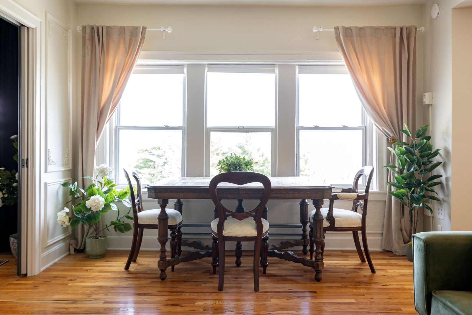 A dining area with a wooden table and chairs, large windows, and a potted plant.