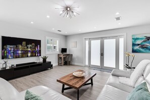 A modern living room with a flat-screen TV, a wooden coffee table, a white sofa, and a large window with blinds.