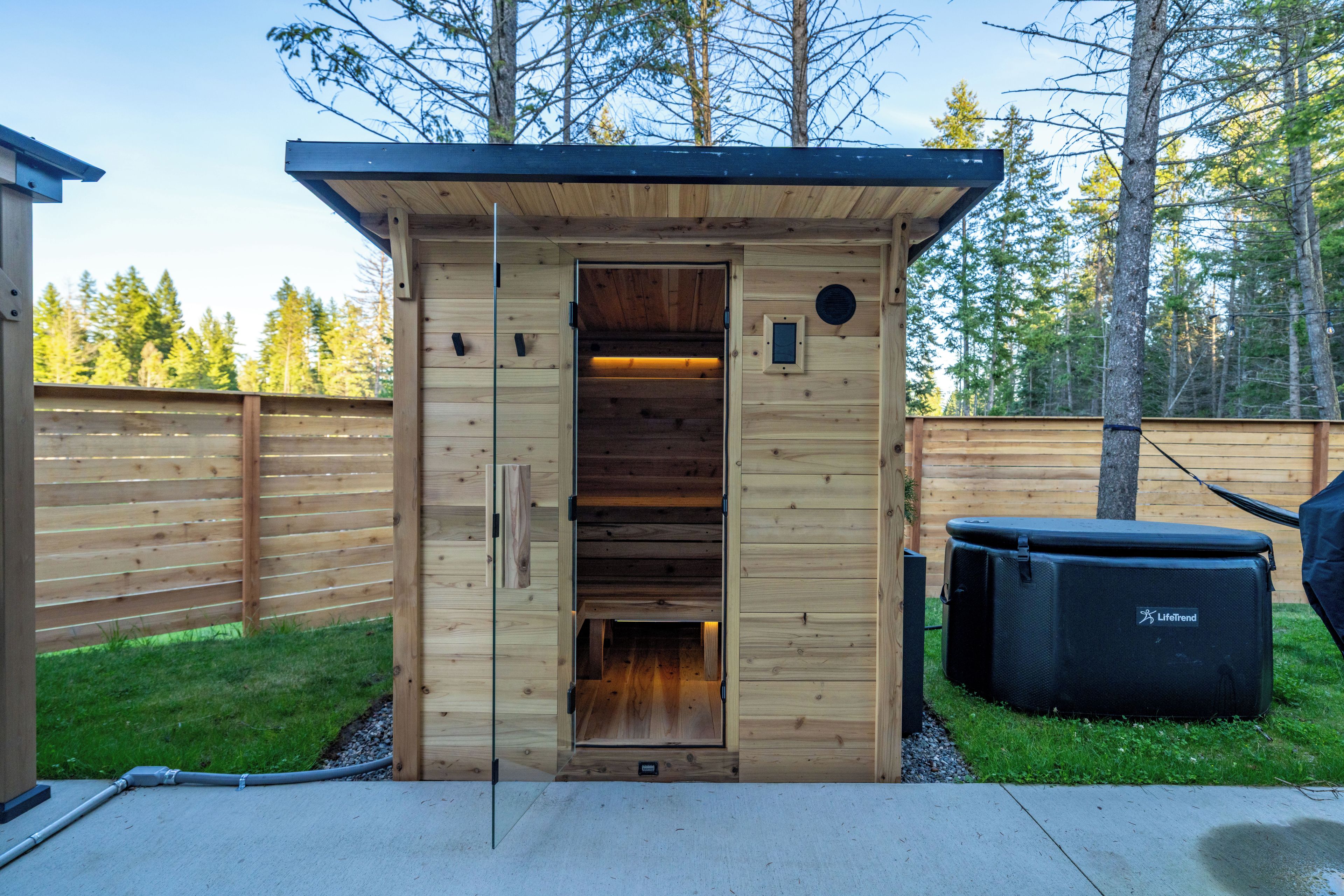 A wooden outdoor sauna with a glass door, a bench inside, and a LifeTrend portable heater outside.