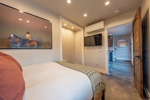 Een slaapkamer met een bed, een wandtelevisie en een gang naar een keuken.