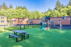 Un’area giochi con scivolo e altalene, delimitata da una recinzione, e un tavolo da picnic su un prato.