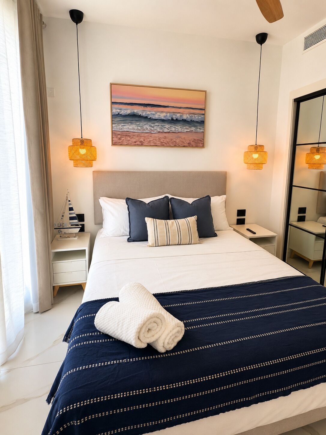 A neatly made bed with a striped blanket, two bedside lamps, a nightstand, and a painting of a beach scene.