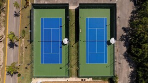 Ein Luftbild eines Tennisplatzkomplexes mit zwei Plätzen, umgeben von Grünflächen und einer Straße.