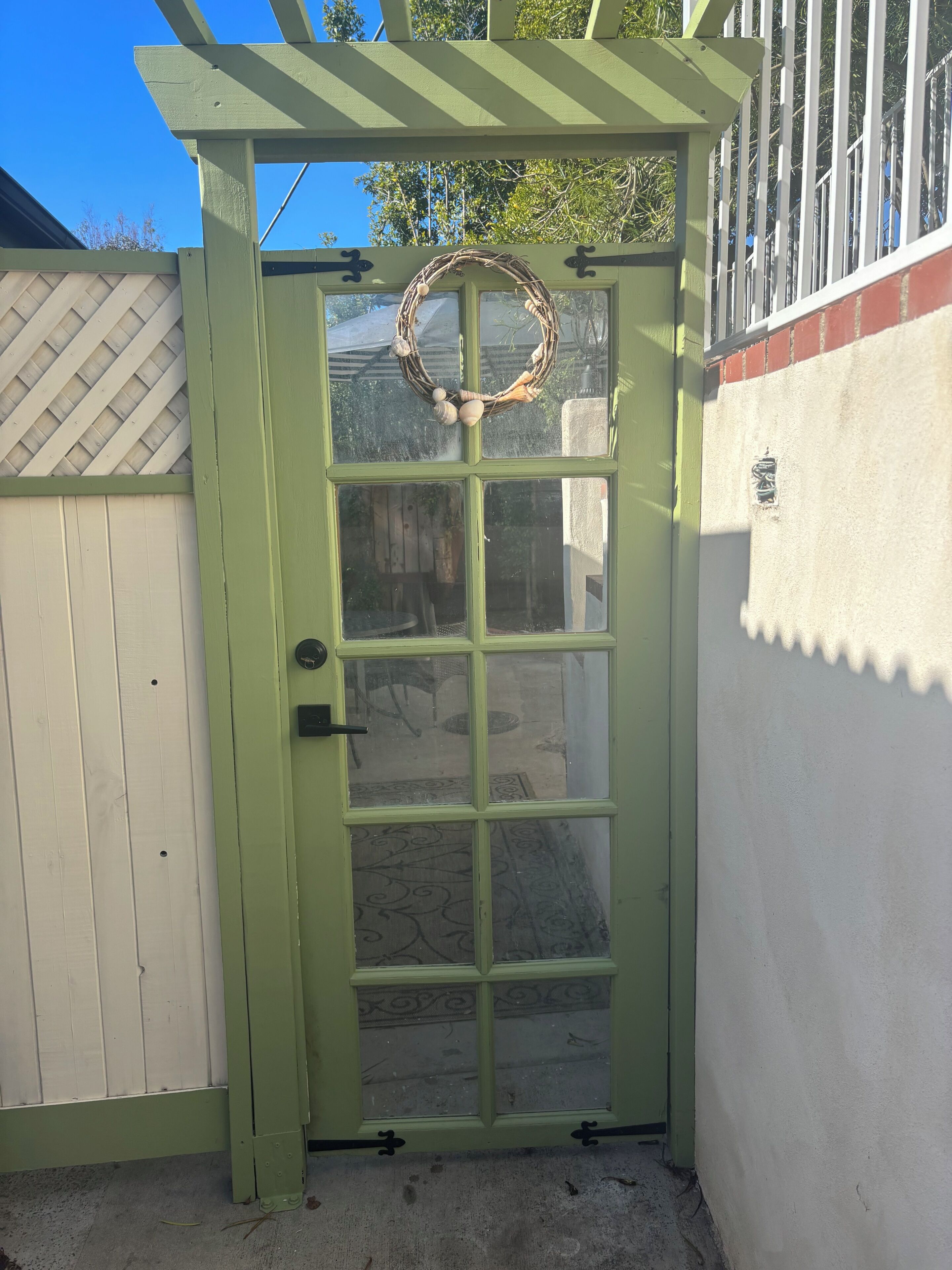 A green door with a decorative wreath, a lattice fence, and a white wall with a textured pattern.