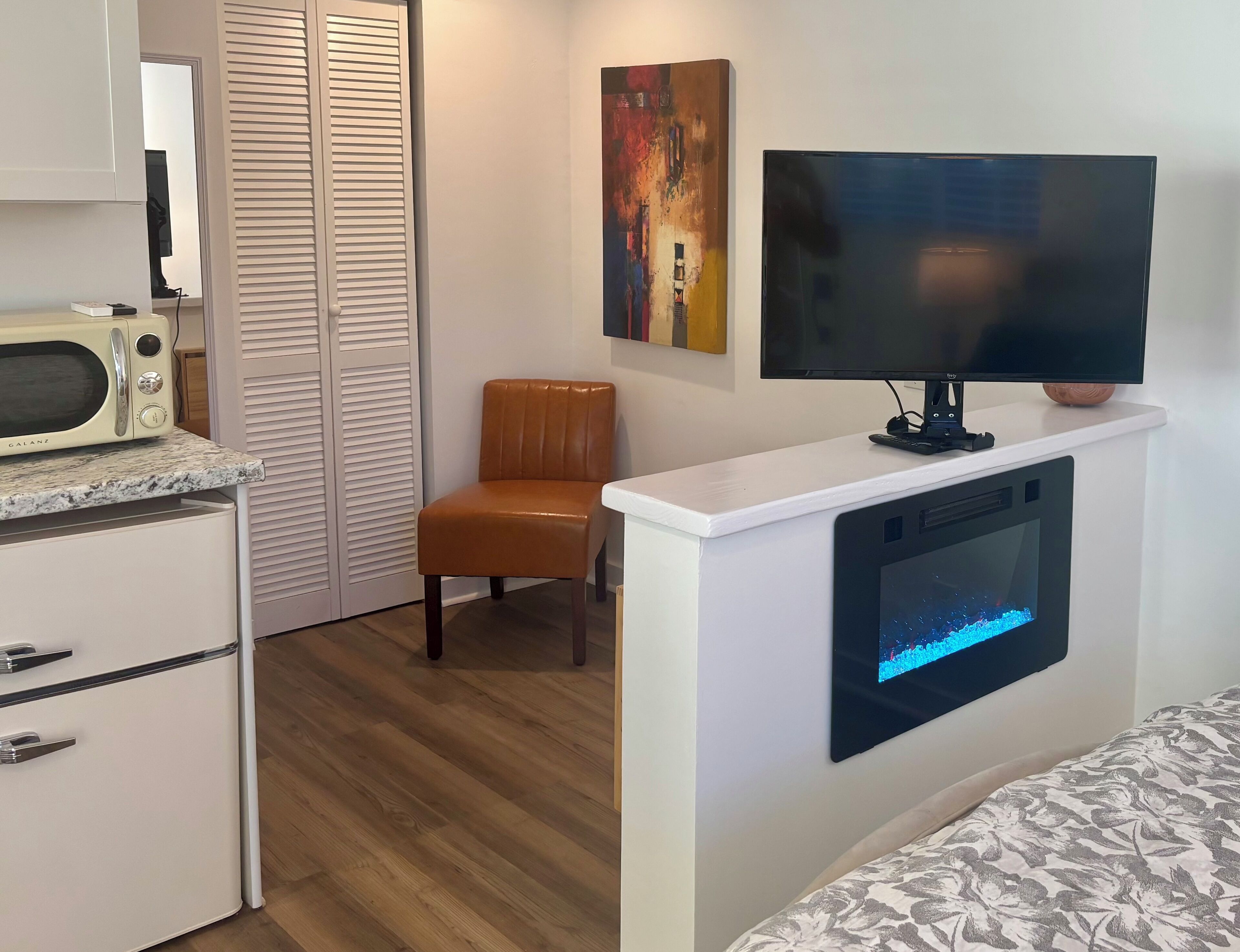 A room with a white cabinet, a microwave, a television mounted on a white wall, a brown leather chair, and a bed with a patterned cover.