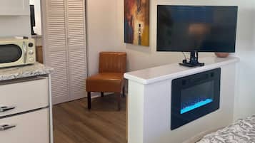 A room with a white cabinet, a microwave, a television mounted on a white wall, a brown leather chair, and a bed with a patterned cover.