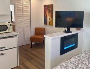 A room with a white cabinet, a microwave, a television mounted on a white wall, a brown leather chair, and a bed with a patterned cover.