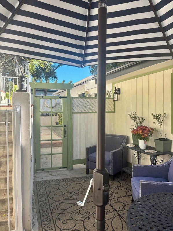 A covered patio area with a striped umbrella, outdoor seating, and a door leading to a building.