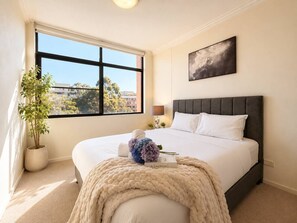 Ein Schlafzimmer mit großem Fenster, einem Bett mit weißen Leinen, einem grauen Kopfteil, einem Nachttisch mit Lampe und einer Topfpflanze.