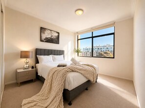 Ein Schlafzimmer mit Bett, Nachttisch, Lampe und einem Fenster mit Blick auf die Stadt.
