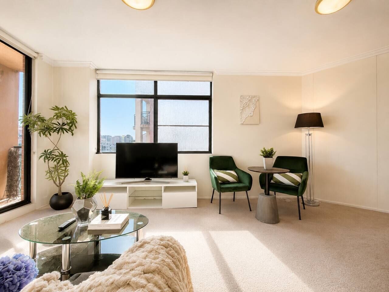 Un salon moderne doté d’un téléviseur à écran plat, de deux fauteuils verts, d’une table basse en verre et d’une grande fenêtre donnant sur la ville.