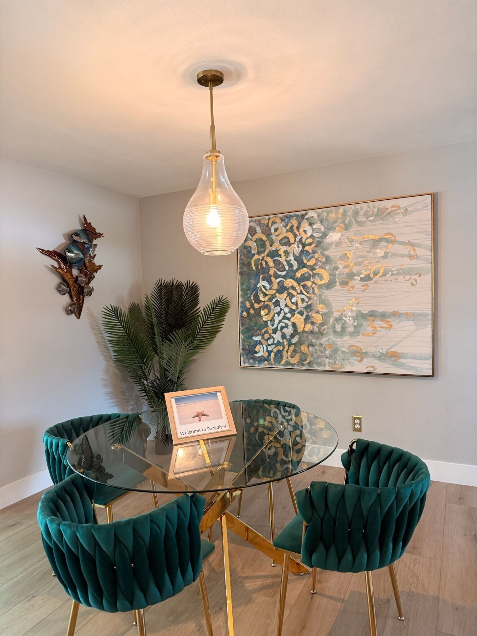 A dining area with a glass table surrounded by teal upholstered chairs, a hanging light fixture, and a framed picture on the wall.