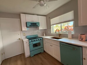 Una cocina con armarios blancos, una estufa verde azulado y una ventana con persianas.