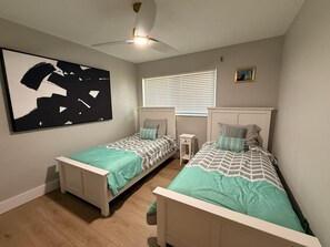 A bedroom with two single beds, a ceiling fan, a window with blinds, and a large abstract painting on the wall. - Retro styled paradise 3bed/2br Cape Coral pool home on the canal front (Cape Coral)