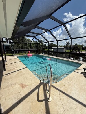 An outdoor swimming pool with a clear blue water, surrounded by a covered area with a metal frame and a view of a residential neighborhood. - Retro styled paradise 3bed/2br Cape Coral pool home on the canal front (Cape Coral)