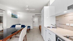A modern kitchen with white cabinets, a stainless steel sink, and a dining area with a table and chairs. - Palm Cove Apartment with Resort Pool, Gym and Parking | 2308 Drift (Palm Cove)