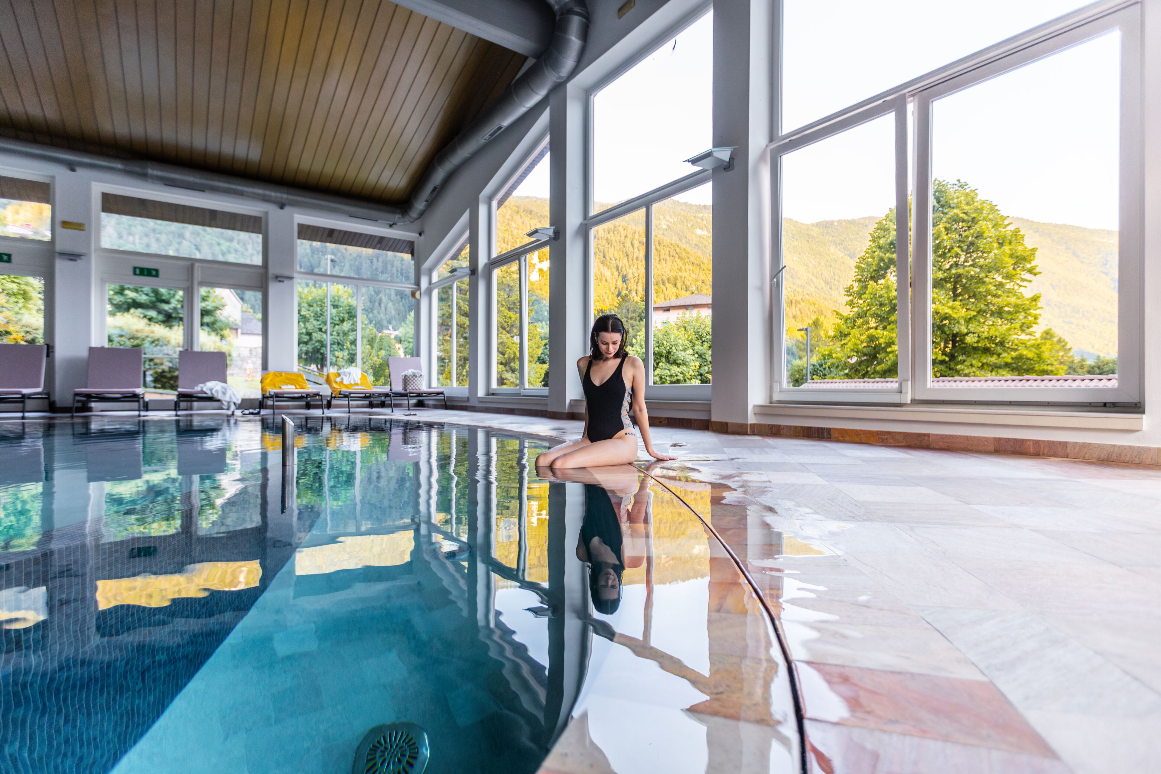 Una persona seduta a bordo piscina, con ampie finestre e vista sugli alberi.