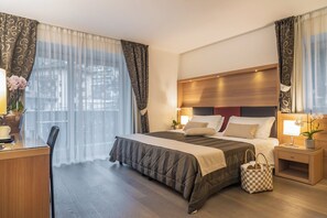 Ein Hotelzimmer mit einem großen Bett, einem Schreibtisch, einem Sessel, einem Nachttisch mit Lampe und Blick ins Freie durch transparente Vorhänge.
