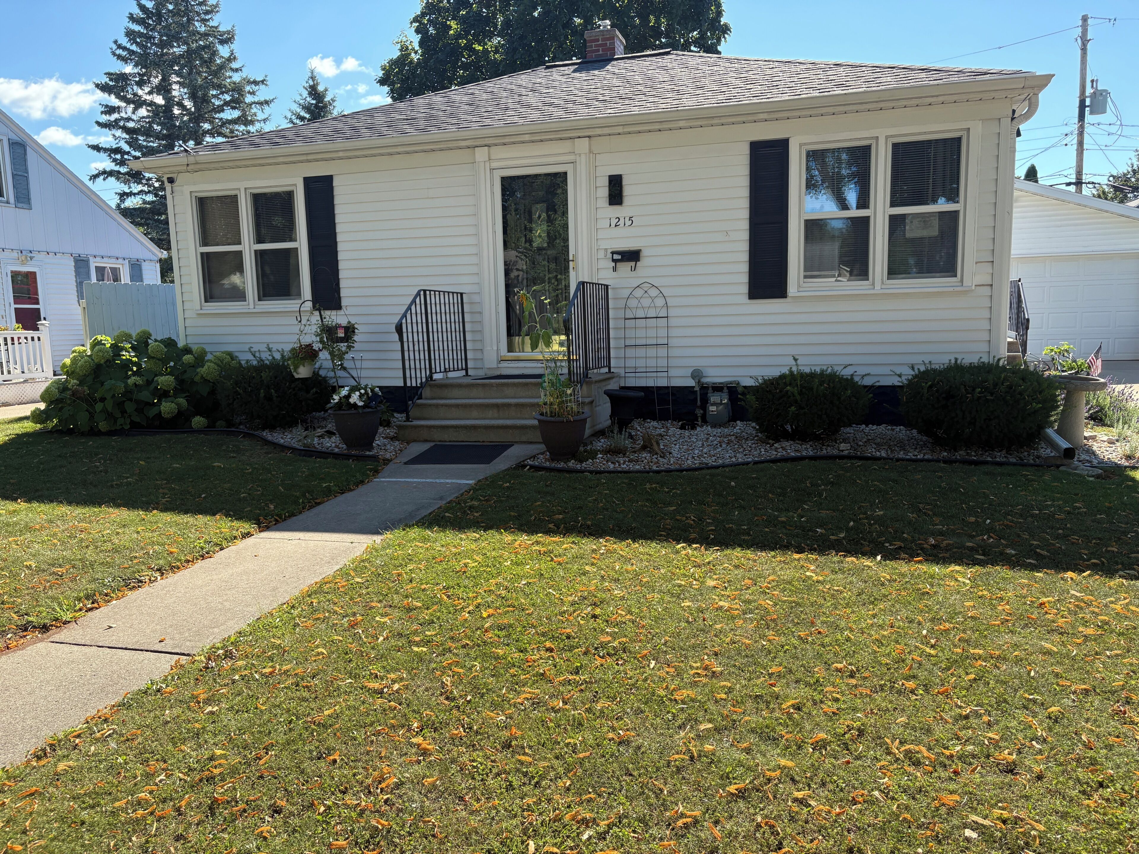 A single-story house with a front porch, a mailbox, and a small garden with shrubs and a potted plant.