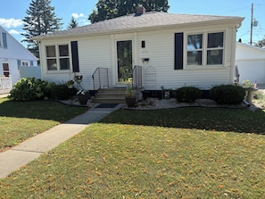 A single-story house with a front porch, a mailbox, and a small garden with shrubs and a potted plant. - Cozy 2-bedroom house in pleasant Green Bay with AC (Green Bay)