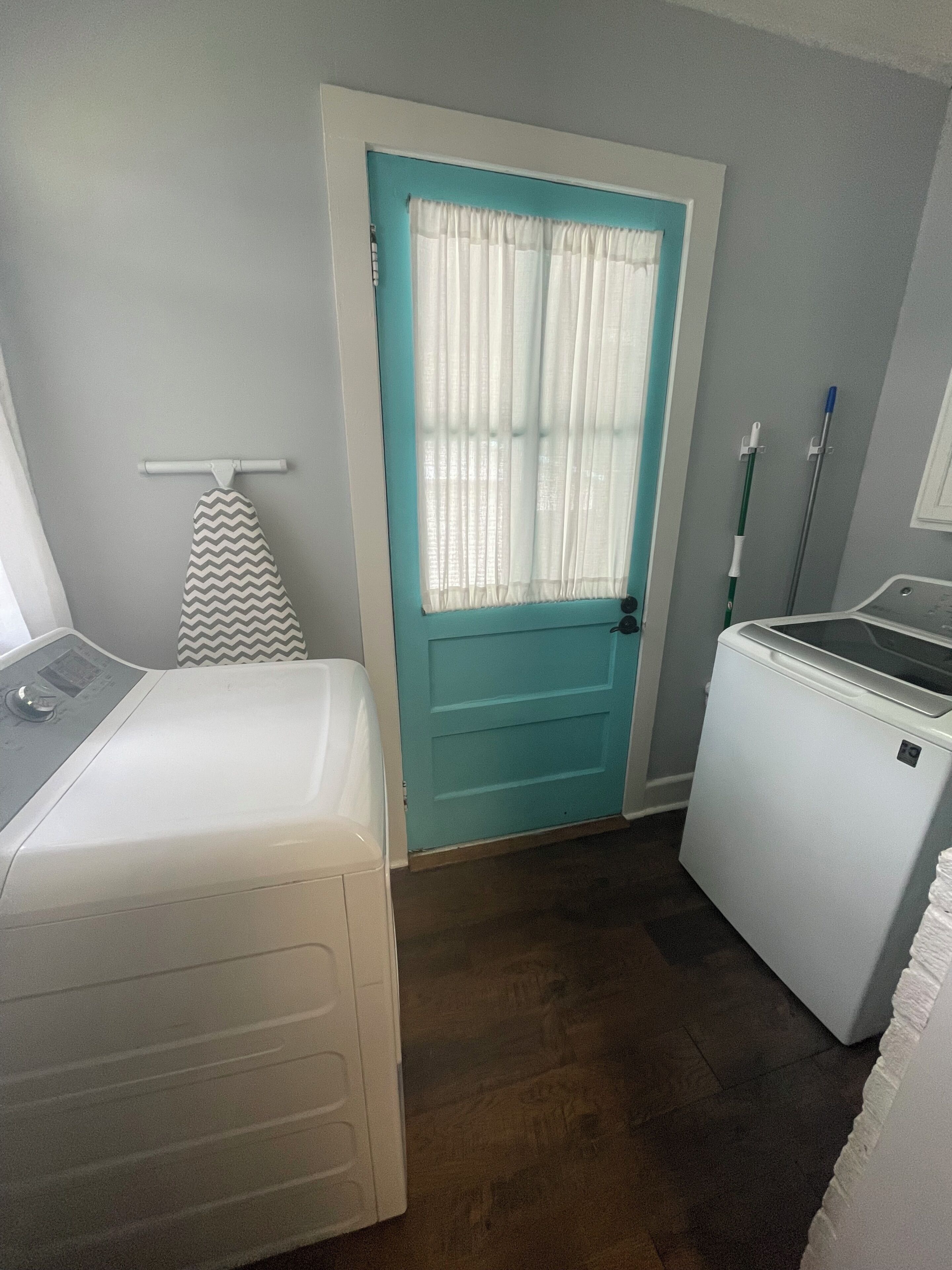 A laundry room with a white washer and dryer, a teal door with a white curtain, and a mop leaning against the wall.
