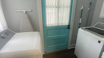 A laundry room with a white washer and dryer, a teal door with a white curtain, and a mop leaning against the wall.