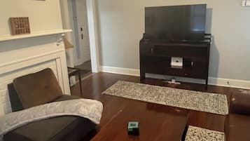 Un salon avec un ventilateur de plafond, un téléviseur posé sur un meuble, une cheminée et une table basse.