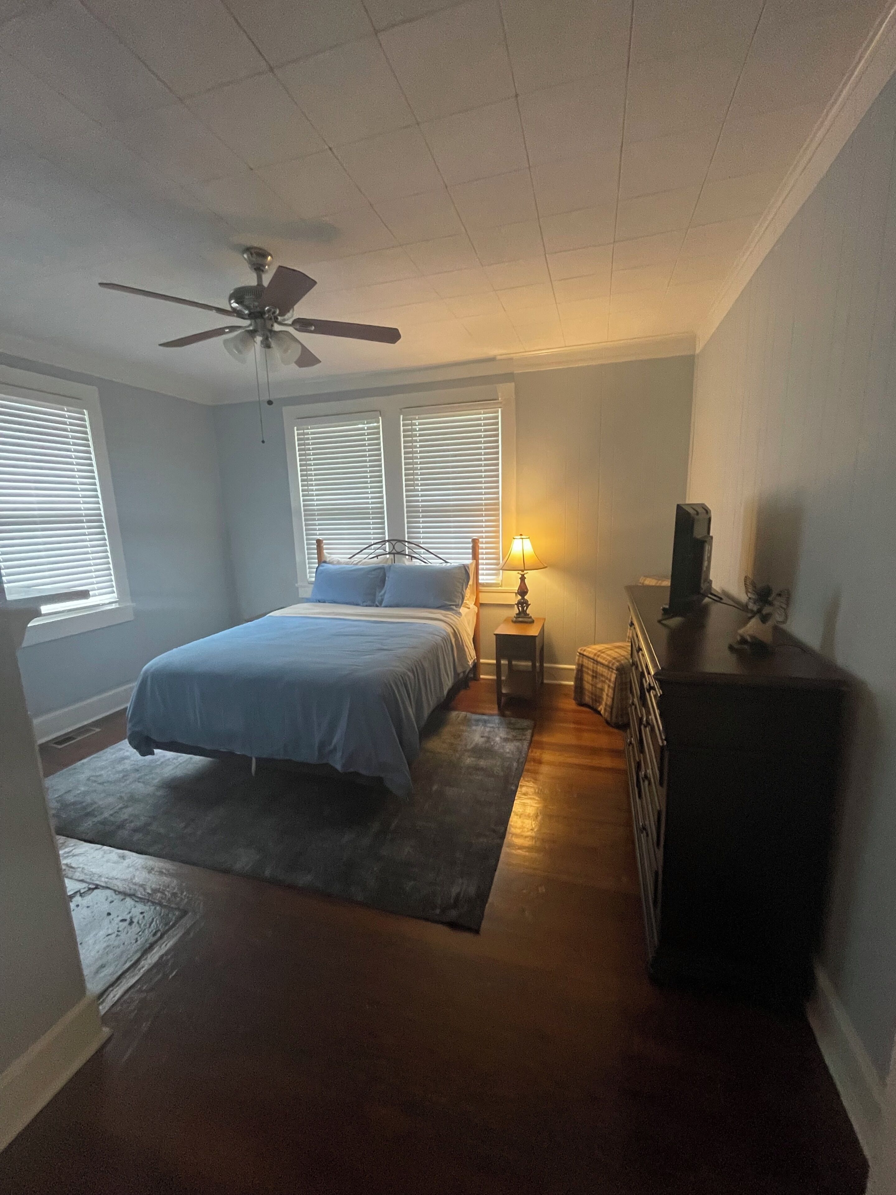 Ein Schlafzimmer mit Bett, Nachttisch, Kommode, Deckenventilator und Fenstern mit Jalousien.