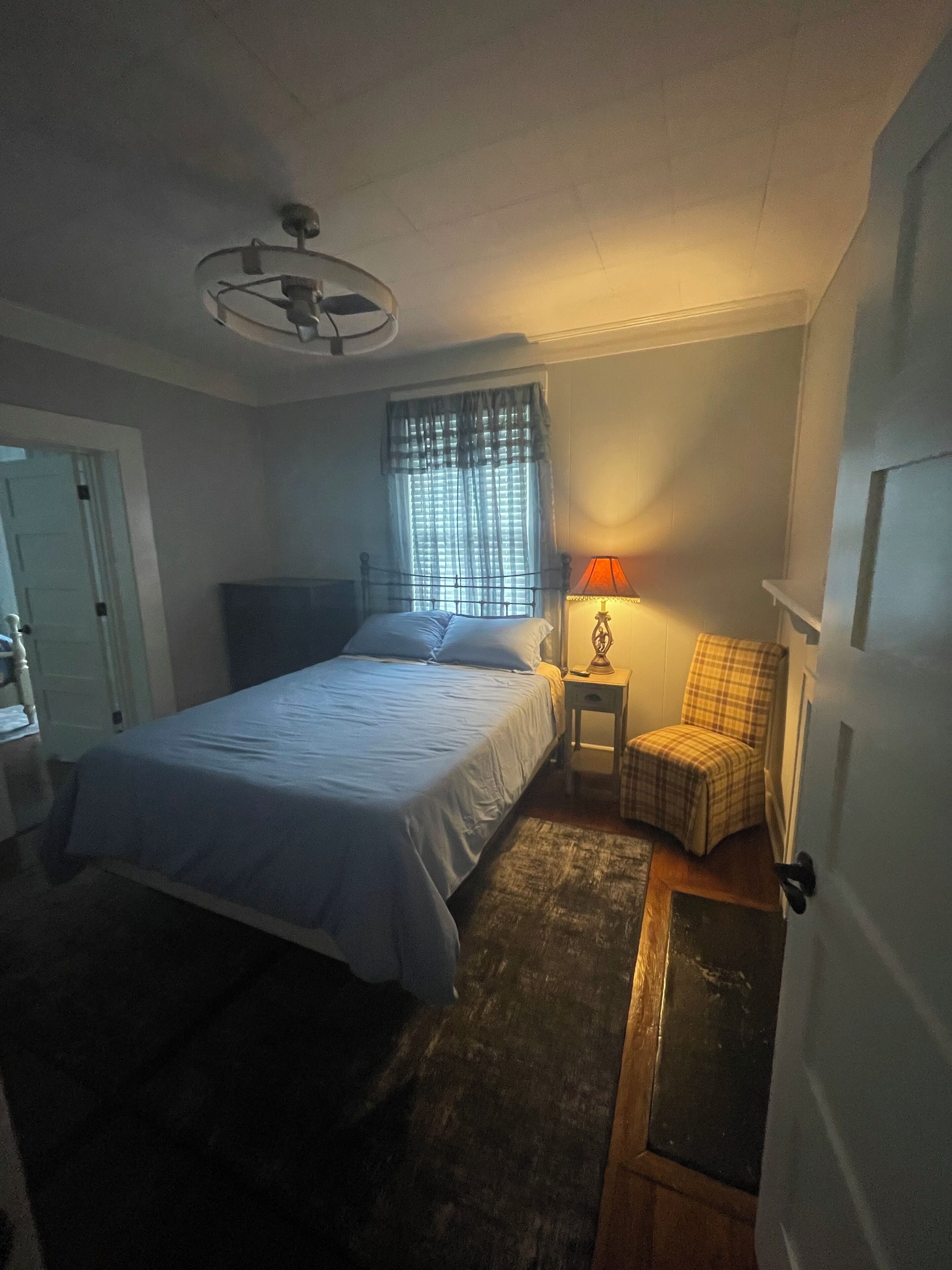 A bedroom with a bed, a nightstand with a lamp, a chair, a window with curtains, and a door.