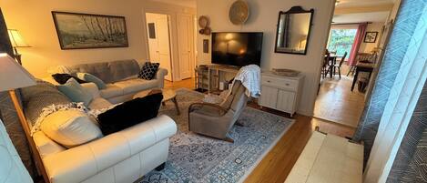 A living room with a sofa, armchair, TV, and a dining area in the background.