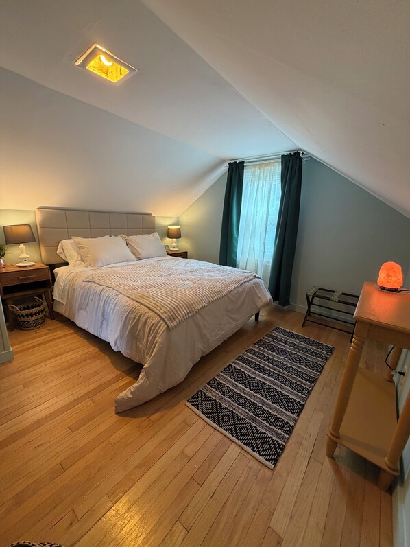 Ein Schlafzimmer mit einem Bett, Nachttischen, einer Lampe, einem Heizkörper, einer Bank und einem Fenster mit Vorhängen.