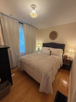 A bedroom with a bed, two bedside tables with lamps, a ceiling light, a window with curtains, and a wall-mounted decorative item. - Cozy home minutes from SU campus, area hospitals & all major highways. (Syracuse)