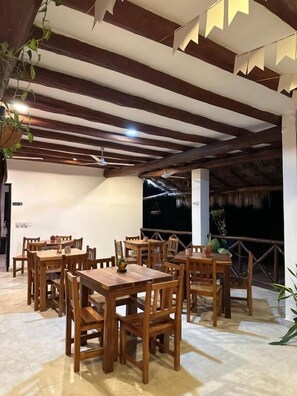 Un comedor interior con mesas y sillas de madera, techo de paja y vistas al cielo nocturno.