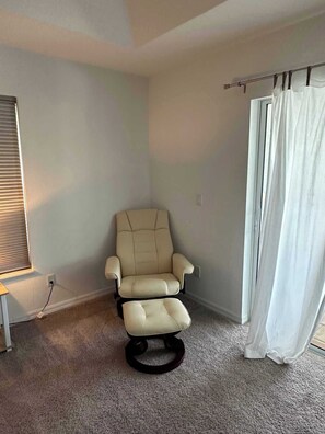 Ein Zimmer mit einem beigen Sessel, einem Fenster mit Jalousien, einem Vorhang und einem Teppichboden.