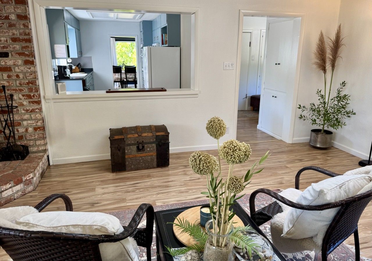 A living room with wicker furniture, a wooden floor, a mirror reflecting the kitchen, and a potted plant.