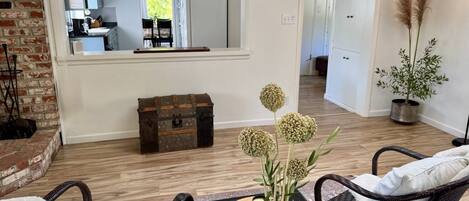 A living room with wicker furniture, a wooden floor, a mirror reflecting the kitchen, and a potted plant.