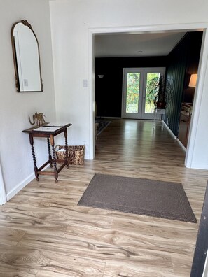 A hallway with wooden flooring, a mirror, a small table with a decorative elephant, and a mat leading to a door with a view of greenery outside.