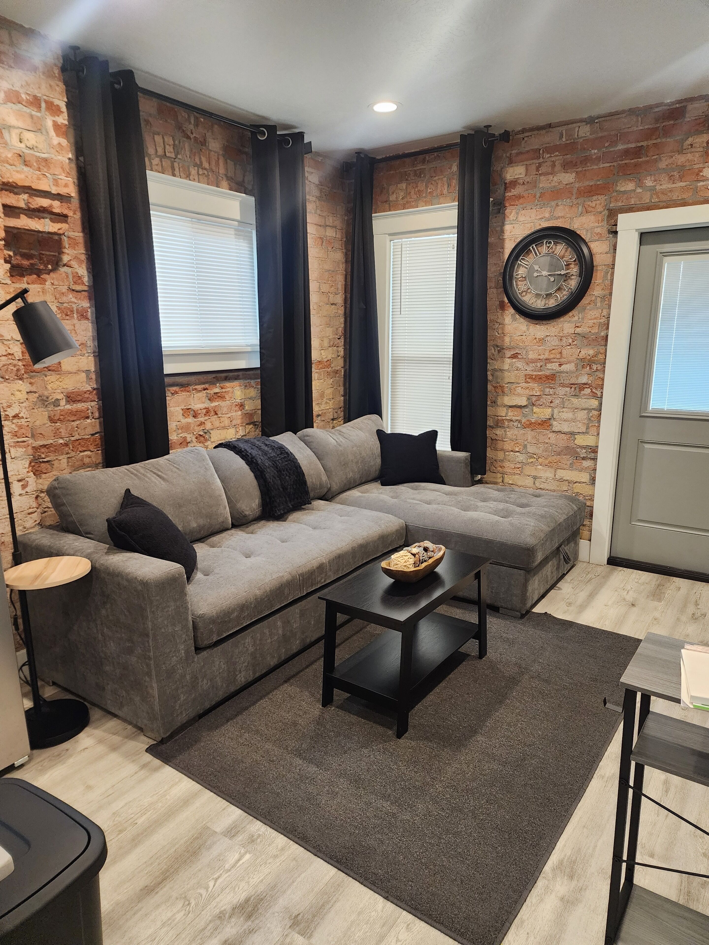 Un salon moderne avec un canapé d’angle gris, une table basse noire et un mur en briques.