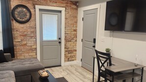 A modern living room with a brick accent wall, a grey sofa, a small table with a bowl, and a TV mounted on the wall. - Cozy 1-bedroom apartment in Beautiful Salt Lake City with W&D (Salt Lake City)