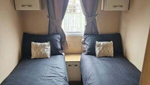A small, enclosed room with two single beds, each with a pillow and a headboard. There is a window with curtains, and a small table or desk is visible in the center.