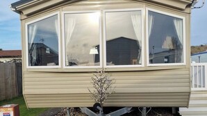 A caravan with a bay window, a small plant in front, and a white fence in the background.