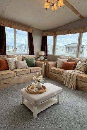 Ein gemütliches Wohnzimmer mit zwei Sofas, einem Couchtisch und einer Lampe. Große Fenster bieten Blick ins Freie.